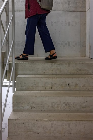 Person in jeans and sandals climbs up a concrete staircase in a building