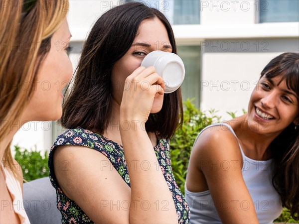 Three women are sitting outside and one of them is drinking from a white cup while looking at camera. They are all smiling and seem to be enjoying each other's company