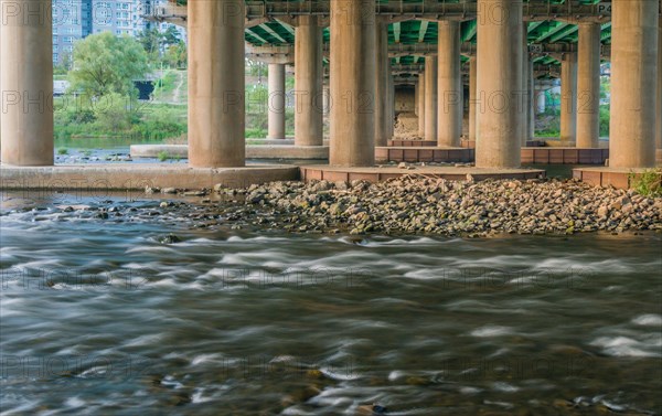 Flowing river under an overpass with concrete columns and greenery around, in South Korea