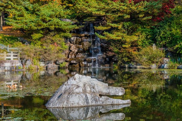 A small waterfall over rocks into a pond, with autumn trees reflecting on the surface, in South Korea