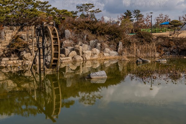 Old water wheel reflected in a pond surrounded by rocks and trees under a cloudy sky, in South Korea
