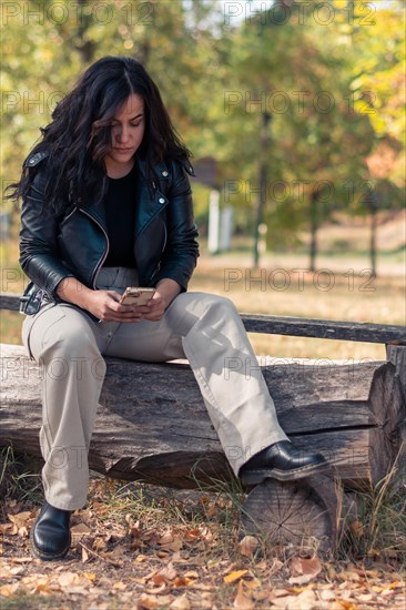 Serious Cheerful hispanic young Woman woman in leather jacket sitting on a bench in a park, looking at mobile phone, in autumn, blurred background with bokeh, daytime, AI generated