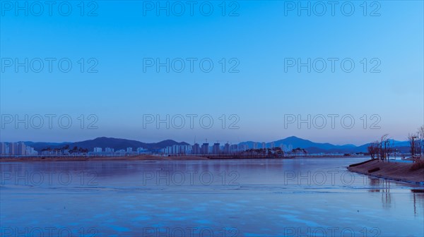Twilight cityscape with a calm river partially covered in ice, reflecting blue hues, in South Korea