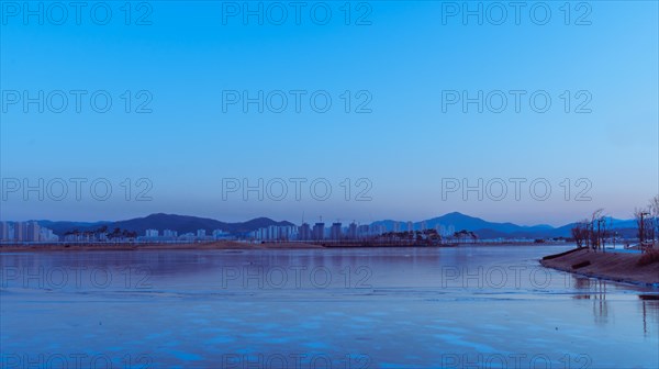 Twilight cityscape with a calm river partially covered in ice, reflecting blue hues, in South Korea