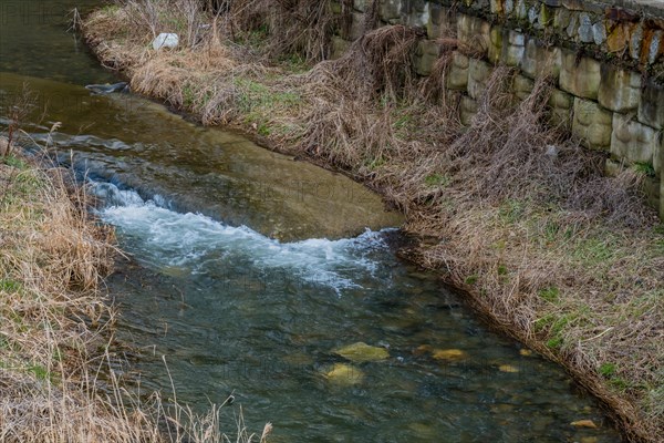 A serene stream flows beside a stone wall lined with dry grass, in South Korea