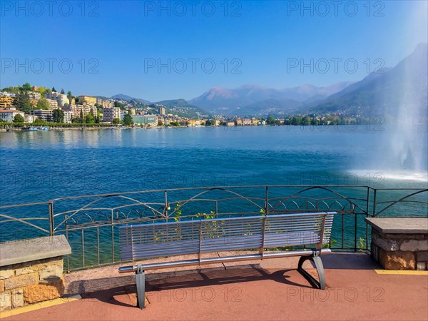 Bench on Lake Lugano and City with Water Fountain and Mountain in a Sunny Summer Day in Lugano, Ticino, Switzerland, Europe