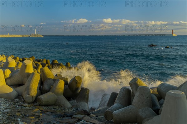 Waves crashing against tetrapods laying on ocean shoreline with two lighthouses on piers on horizon under partly cloudy morning sky, in South Korea