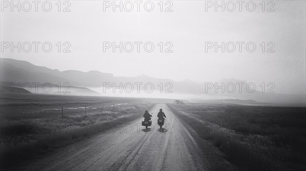Two motorcyclists ride down a misty dirt road with mountain silhouettes in the distance, AI generated