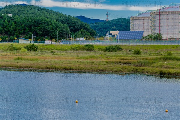 Industrial storage tanks beside a calm water body dotted with buoys, set against a mountain backdrop, in South Korea
