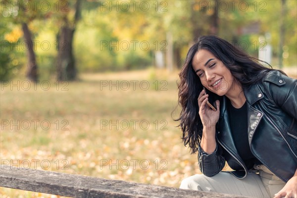 Hispanic Woman smiling during a relaxed phone conversation with mobile phone, sitting on a wooden bench outdoors, outdoors in autumn, blurred background with bokeh, daytime, AI generated