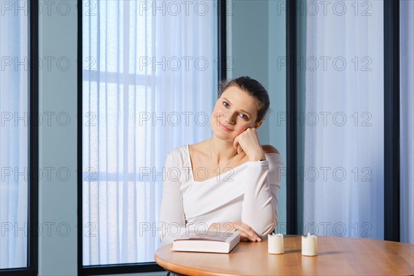 Dreamy woman sits at a wooden table with a book, props her head up with her fist and looks with a kind smile