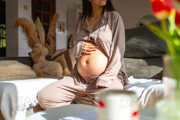 An unrecognizable pregnant woman touching her belly wearing pajamas sitting on the sofa