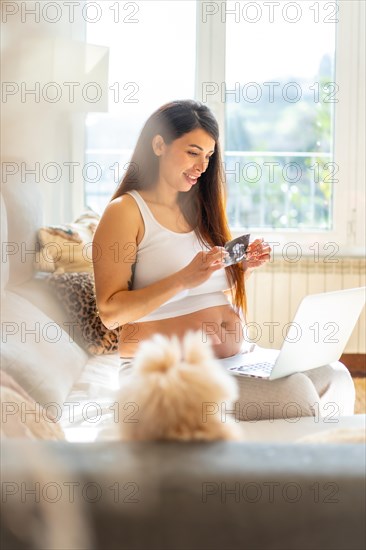 Vertical photo of a breauty adult pregnant woman showing baby ultrasound in video call using laptop sitting on the sofa at home