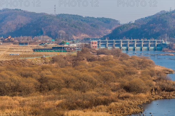 River cutting through a landscape with dam, houses, and hills under a clear sky, in South Korea