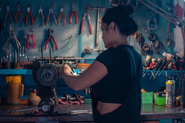 Skilled craftswoman repairing a motorcycle engine part in a workshop with a teal wall, a complete tool panel in background with bokeh effect, traditional male jobs by Mixed-race latino woman