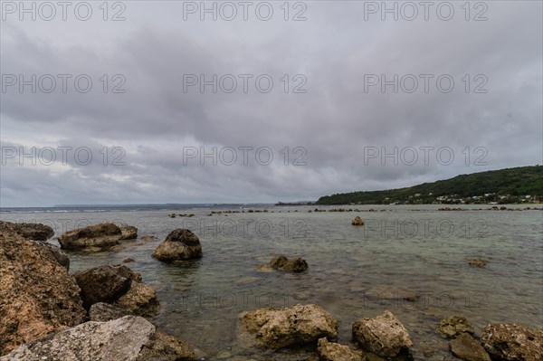 Seascape of rocky shoreline on a cloudy day with buildings on tree lined shore in the distance in Guam
