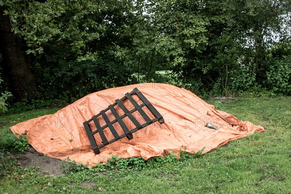An orange tarpaulin covers something on the ground next to a wooden pallet and bushes