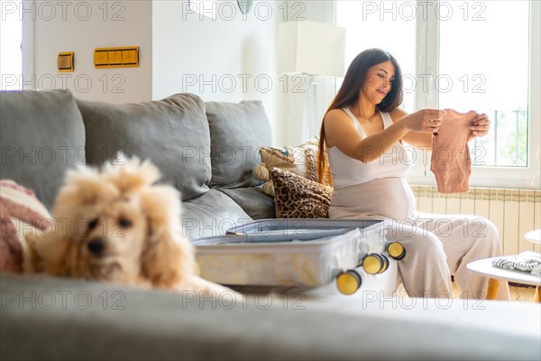 Happy pregnant woman getting ready to go to the hospital preparing clothes and suitcase sitting on the sofa next to a dog