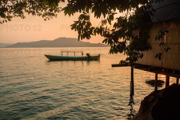 A solitary boat glides on tranquil water under a sunset sky near a silhouette of a tree. Koh Sdach Island, Cambodia, Asia