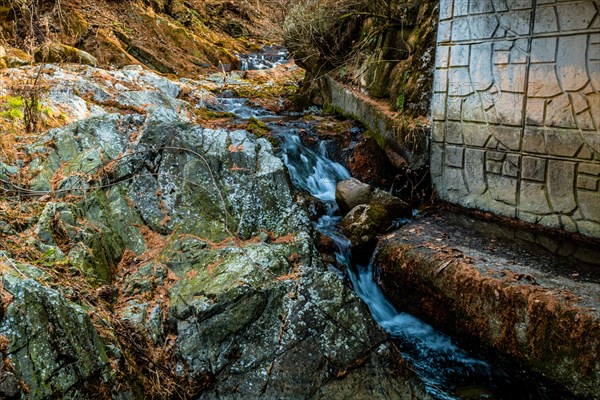 Water cascades over rocks in a stream next to a brick wall in a natural wooded landscape, in South Korea