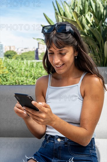 A woman is sitting on a couch and looking at her cell phone. She is smiling and she is happy