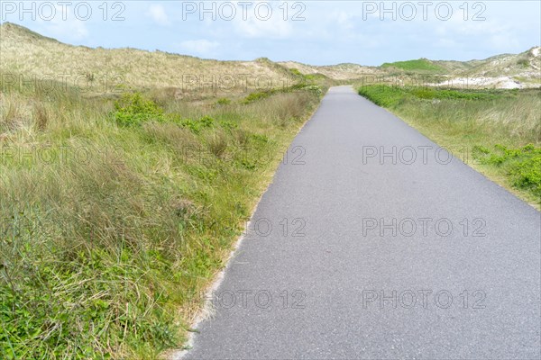 A straight tarmac road leads through a wide dune landscape under a cloudy sky