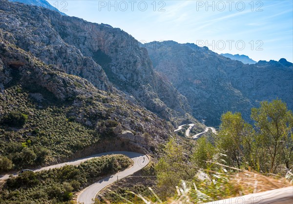 Mountain landscape with winding mountain road from Sa Calobra to Torrent de Pareis, cars and a cyclist on serpentines with various hairpin bends, hairpin bends, hairpin bends, steep, partly bare rock faces and peaks, Mediterranean vegetation, power line, trees and grass, Serra de Tramuntana mountains, sunny weather and blue sky, panoramic road, Majorca, Spain, Europe
