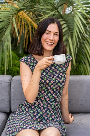 A woman is sitting on a couch and holding a cup of coffee. She is smiling and she is enjoying her drink ahile looking at camera