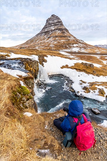 Adventurous photographer woman in winter in Iceland photographing sitting at the Kirkjufell waterfall and its beautiful mountain
