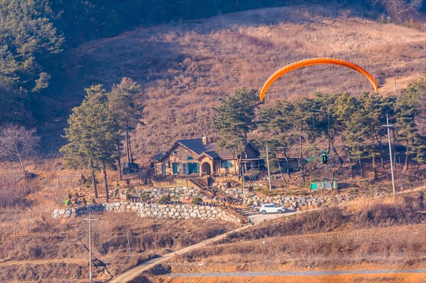 Paraglider above a house surrounded by trees in a rural landscape, in South Korea