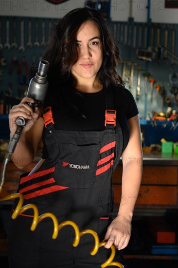 A confident woman mechanic with a tool in a workshop wearing overalls, a complete tool panel in background with bokeh effect, traditional male jobs by Mixed-race latino woman