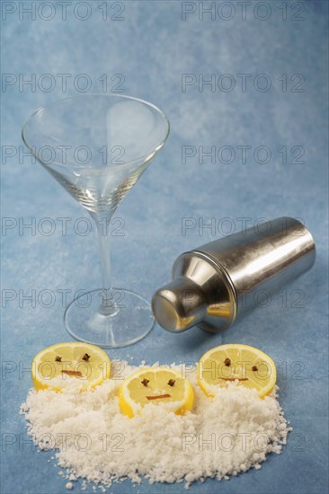 Lemon slices with cloves and cinnamon forming a smiley face on a pile of salt next to a cocktail shaker and a crystal glass with blue background with copy space