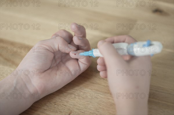 On the left a child's hand with a drop of blood on the little finger, in the right hand the child holds the lancet, lancing device, has just pricked himself, wooden table as background, blood glucose measurement, diabetes treatment, glucose measurement, Ruhr area, Germany, Europe