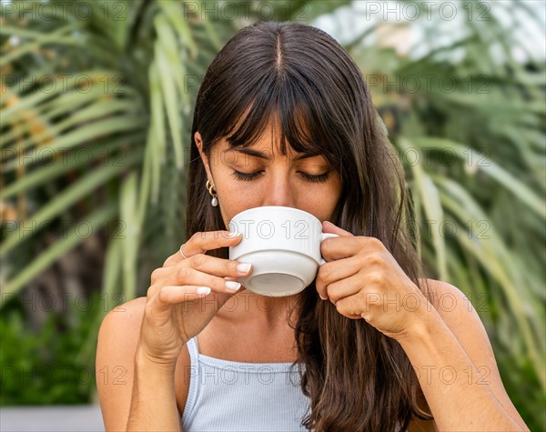 A woman is drinking coffee from a white cup with her eyes closed. She is wearing a tank top and has her hair pulled back