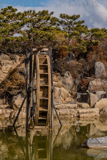 Close-up of a water wheel with its reflection in a pond, set against a rocky backdrop, in South Korea