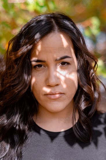 Close-up of a Cheerful hispanic young woman with a serious expression and direct gaze, selective focus, blurred background with bokeh, daytime, AI generated