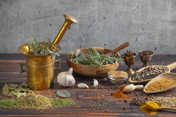 Brass mortar with herbs surrounded by spices and kitchen utensils on wood