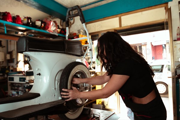 Hispanic young sensual long haired brunette woman Mechanic adjusting a tire on a vintage italian scooter at a workbench, latino female in traditional masculine jobs concept, feminine power in real life