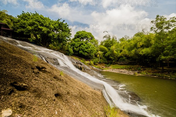 Side view of a waterfall cascading down a cliff into the basin below with green trees and blue, cloudy sky in the background in Guam