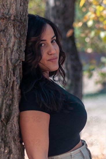 A relaxed Cheerful hispanic young latina sensual woman leaning against a tree trunk amidst nature, casually dressed in a black shirt, selective focus, blurred background with bokeh, daytime, AI generated