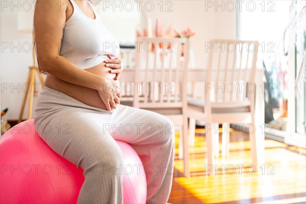 Unrecognizable pregnant woman touching her belly during an active pregnancy at home doing exercises with pilates pink ball