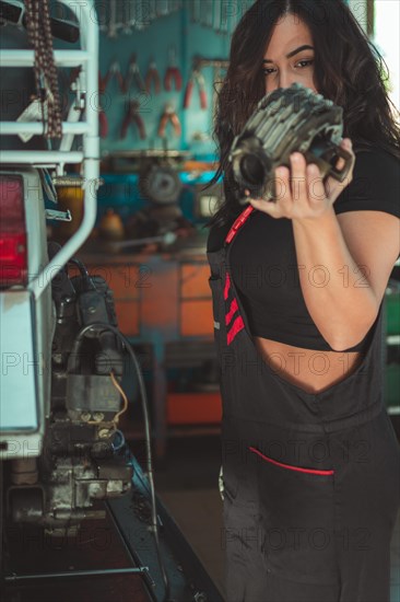 Professional latino female motorbike mechanic holding component of italianvintage scooter in a workshop, a complete tool panel in background with bokeh effect, traditional male jobs by Mixed-race hispanic woman