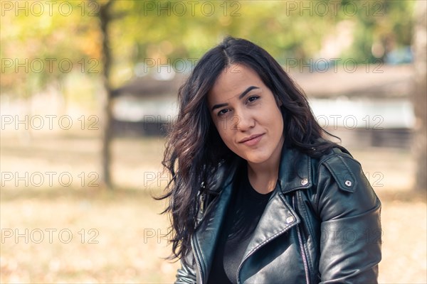 A confident hipanic woman in a black leather rocker jacket smiling outdoors in autumn, blurred background with bokeh, daytime, AI generated