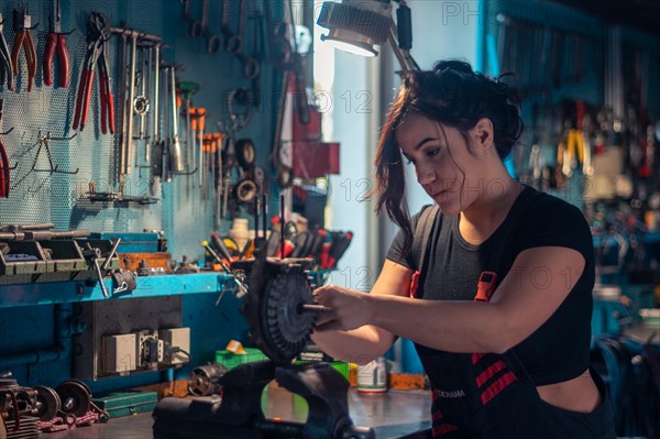 Serious young mechanic concentrating on repairing a scooter engine part in a vise in a well-lit workshop, a complete tool panel in background with bokeh effect, traditional male jobs by Mixed-race latino woman