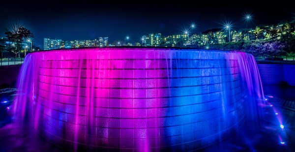 Night scene of waterfall lit up with blue and red lights with the lights of buildings of a town in South Korea in the background in Sejeong, South Korea, Asia