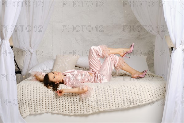 Playful woman in pink satin pajamas and stilettos lying on her back on bed in cozy bedroom