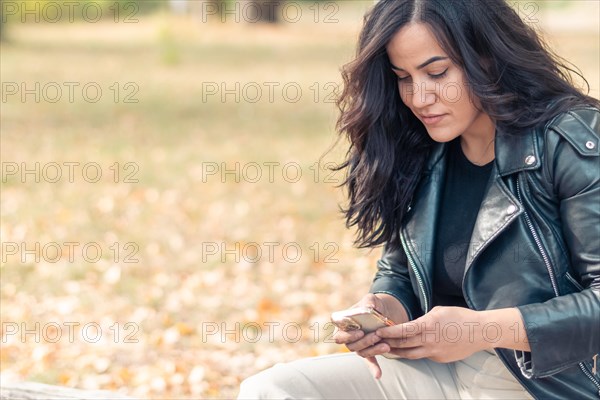 Latino Woman sitting on a park bench focused on her smartphone, wearing casual clothes, outdoors in autumn, blurred background with bokeh, daytime, AI generated