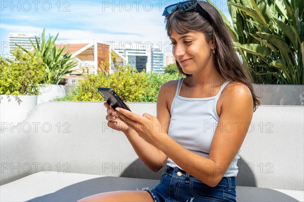 A woman is sitting on a couch and looking at her cell phone. She is wearing a white tank top and blue jeans
