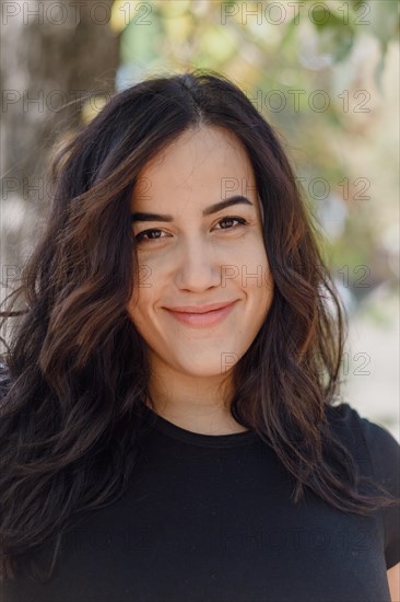 Outdoor image of a confident Cheerful hispanic young woman smiling in a black shirt with a relaxed and pleasant demeanor, selective focus, blurred background with bokeh, daytime, AI generated