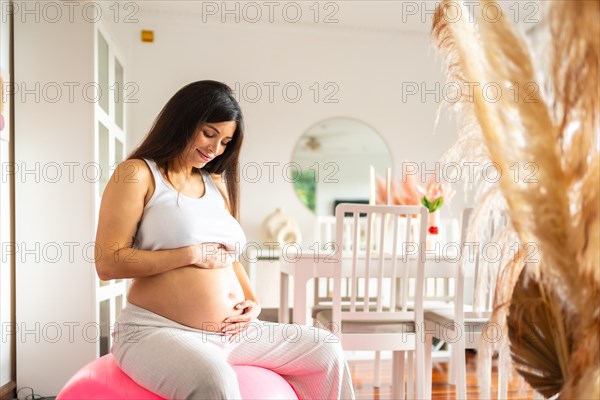 Sportive beauty pregnant woman touching her belly while sitting on pink pilates ball at home